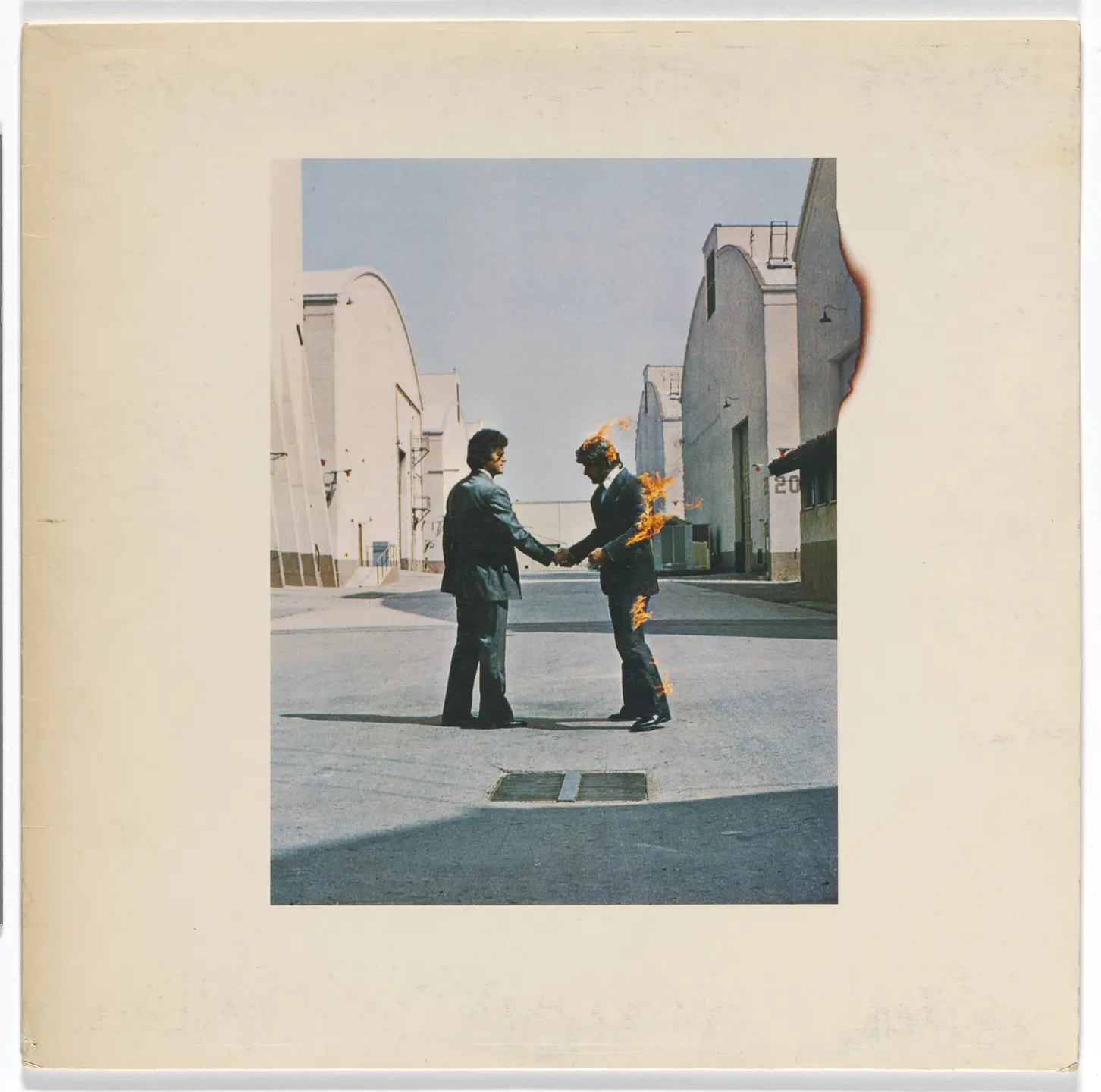 The album cover of Wish You Were Here (1975) shows two men in business suits shaking hands in a film studio backlot. One of the men is on fire, engulfed in flames, while the other appears unaffected. The scene is staged against an industrial Hollywood-style setting with sound stages and clear blue sky.